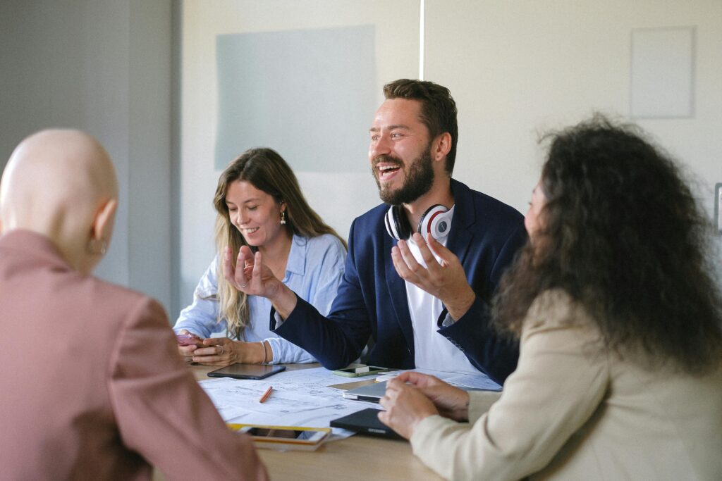 Image shows four people sat around a table having a conversation. Two people have their back to us and are both wearing suit jackets, one on the right and one on the left. Two people in the centre are facing us. The one on the left is looking down and smiling. The one on the right is gesturing with his hands facing upwards, and talking with an animated expression. The implication is that people are working together in a constructive way.