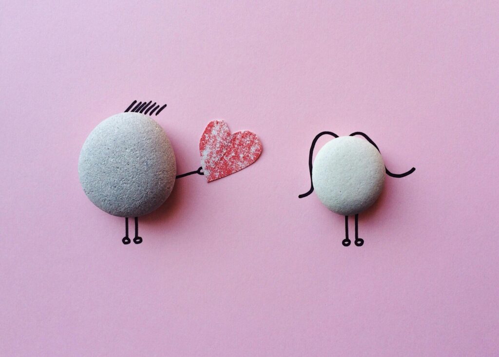 Image shows two rocks on a sheet of pink paper. Someone has drawn on the paper behind each rock to give it legs and hair. The rock on the left has spiky hair and two legs. It has an arm holding out a red paper cut out heart towards the rock on the right. The rock on the right has two legs and two long wavy lines for hair.